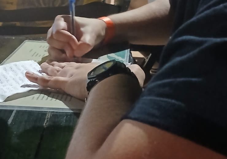 As mãos de Anderson Nunes escrevendo uma letra à caneta num pequeno papel, sobre uma mesa.